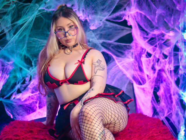 DandelionQueen with big boobs, wearing black red-trimmed lingerie, fishnet stockings, and glasses, has tattoos, a choker, and long blonde hair, sitting on a red fluffy surface.