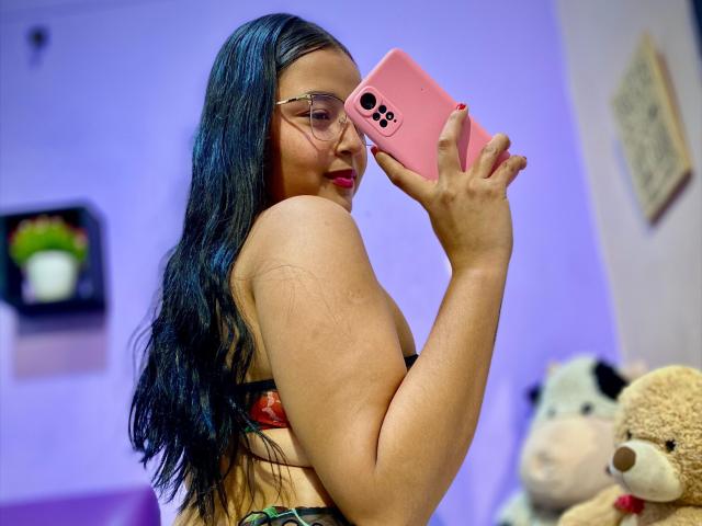 YisethLucia with long black hair, glasses, pink phone, and green bra, taking a selfie, showing her back and round ass, in a bedroom with stuffed toys.