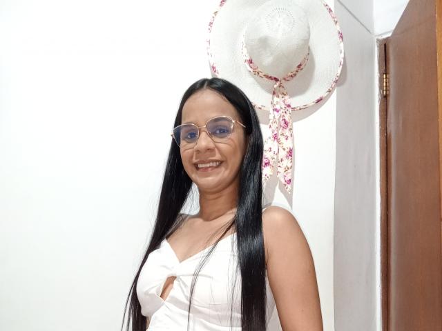 YisethLucia with long black hair, glasses, smiling, wearing a white tank top showing small tits, standing next to a floral sunhat on a white wall.