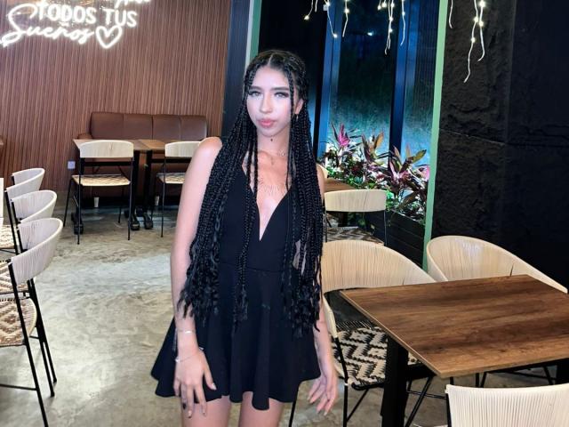 AriRosse with long black braids, wearing a black dress, standing in a restaurant, showing her tits and ass, looking at the camera with a slight smile.