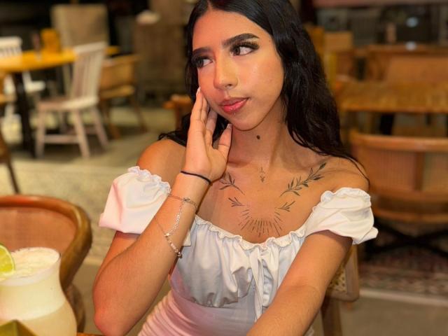 AriRosse with long black hair, pouty pink lips, and tattoos under her tits, wearing a white off-shoulder top, touching her cheek in a cozy restaurant.