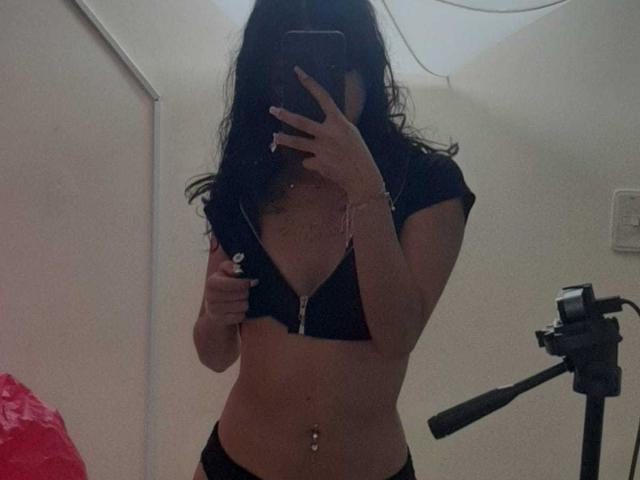 AriRosse with wavy black hair, holding a phone, wearing a black bra and unzipped top, showing her tits and belly button, mirror selfie, camera on right.