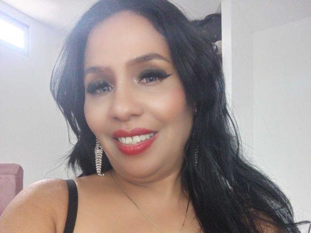 ZuryMasson has long black hair, red lipstick, and is smiling with big eyes and eyeliner. She's wearing a black top and diamond earrings, with a visible tan.