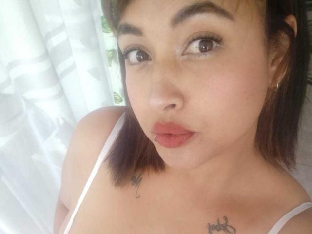 MirandaDreams has light brown skin, brown eyes, and a small nose piercing. She's wearing a white top, showing small tits, and has tattoos on her chest. Her lips are puckered.