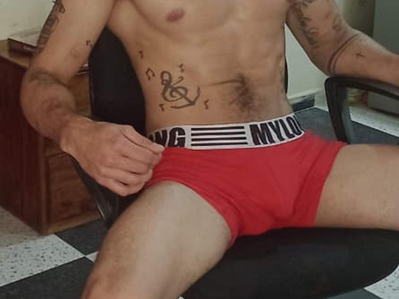 JeffersLHom with a toned, tattooed torso, wearing red boxer shorts, sitting on a chair, right hand near waist, showing a small, flaccid bulge.