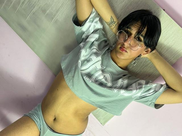NeonAdonis with short black hair, glasses, and a tattoo on the upper arm, wearing a gray crop top and matching underwear, lying down with arms behind head.