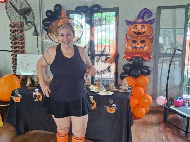 LadySelene, a chubby woman with big tits, wearing a witch hat, black tank top, and black skirt, stands in a Halloween-themed room with orange and black balloons.