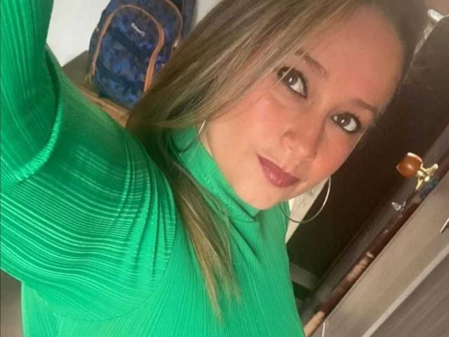 BrendaBigTitis with long blonde hair, green ribbed turtleneck, and hoop earrings, smiling at the camera, holding up her phone for a selfie. Her tits are subtly visible.