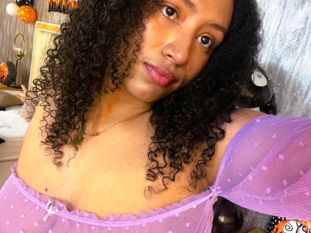 KarinaLovey with curly black hair, brown eyes, and pink lipstick, wearing a purple sheer top that reveals her tits. She has a gold necklace and a subtle smile.