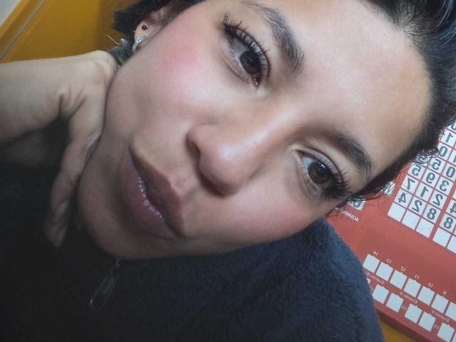 PaulinaColemann's face, slightly tilted, with glossy lips, dark eyes, and a hand near her neck, wearing a black top, showing a cute, pensive expression.