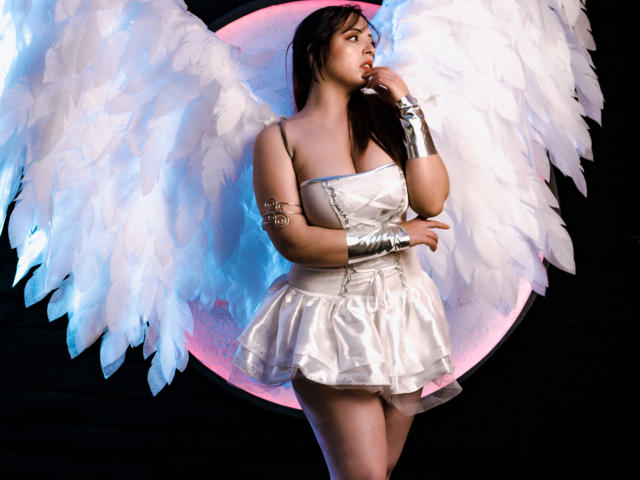 Rosalinnes with white angel wings, wearing a shiny white dress and silver arm cuffs, has a tattoo on her right arm, posing with her finger to her lips.
