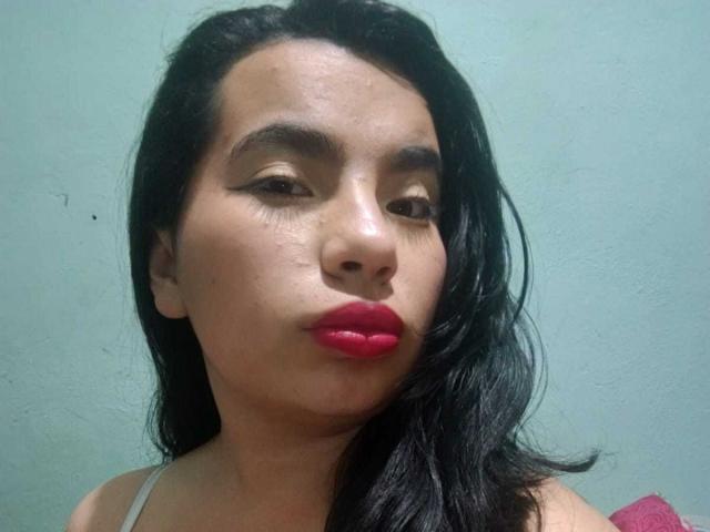 MaayGrey has dark, wavy hair, tan skin, and is pouting with bright red lipstick. She's wearing a grey top and has a small, visible nose bump.
