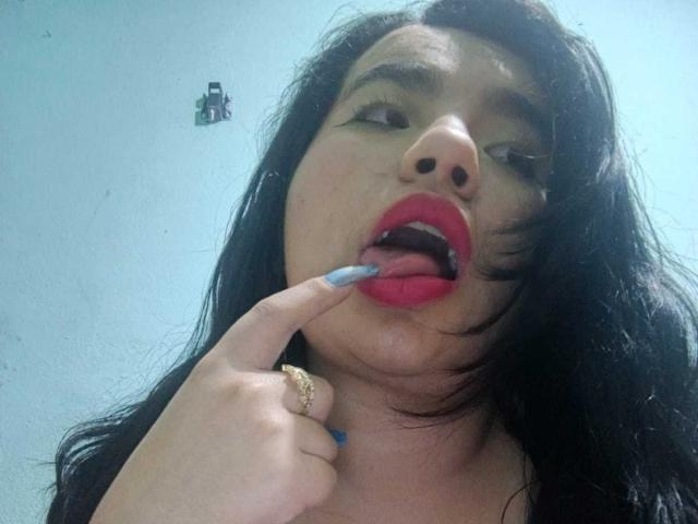 MaayGrey with black hair, red lipstick, and blue nail polish, sucking on her finger, mouth open, showing teeth, wearing a gold ring, looking up, light skin, slight blush.