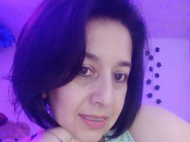 Cami69s has pale skin, dark brown wavy hair, and brown eyes with a slight smile, showing pink lips. Purple light casts a soft glow on her face.