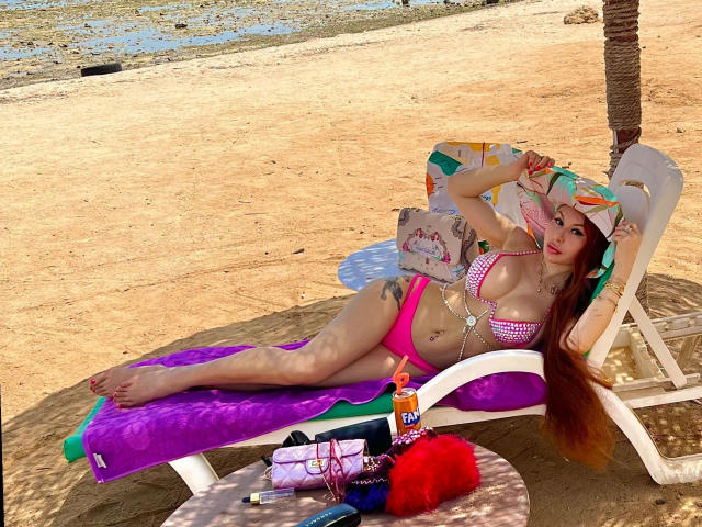Catlady-hot lounging on a purple towel, wearing a pink and white checkered bikini, with long brown hair, and a sun hat, beside a beach chair with sunscreen and a bag.