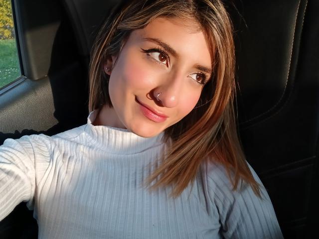 Kaiix a les cheveux bruns clairs, un anneau au nez et un sourire charmant. Elle porte un pull blanc côtelé à col roulé, est assise dans une voiture, et le soleil brille sur son visage..