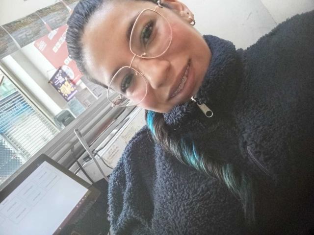 Paulina Colemann mit Brille, einem schwarzen, flauschigen Pullover und einem blau-grünen Zopf, lächelt in die Kamera. Ihre Brüste sind klein, und sie trägt ein Kleidungsstück mit Reißverschluss. Ihr Hintern ist nicht sichtbar..
