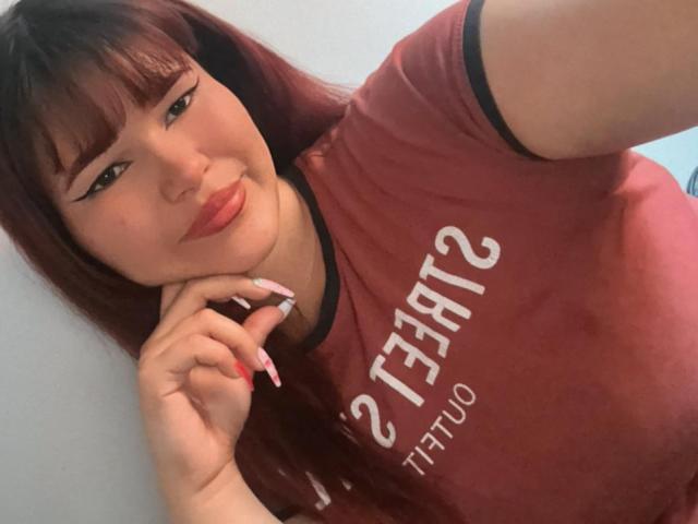 DaliaNova, avec ses cheveux roux, sa frange et son rouge à lèvres rose, portant une chemise bordeaux, avec un sourire en coin, touchant son menton avec ses doigts manucurés en rose, regardant l'appareil photo. Seins, poitrine, mignonne..