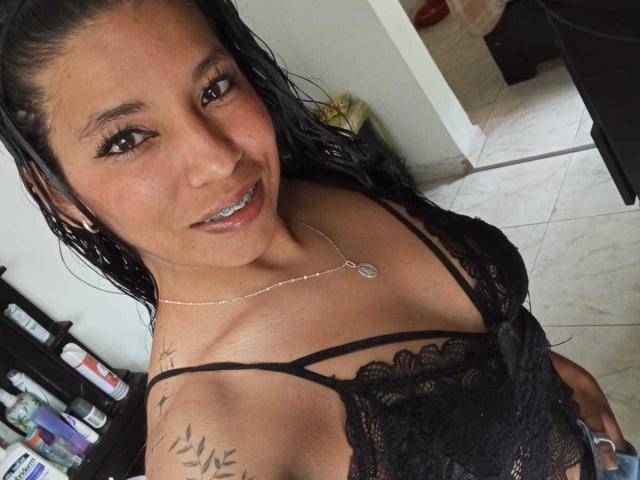 Paulina Colemann mit nassen, schwarzen Haaren, trägt eine schwarze Spitzenbluse, die ihre Brüste zeigt, hat ein Tattoo an der Schulter, eine silberne Halskette, lächelt und steht in einem gefliesten Raum..