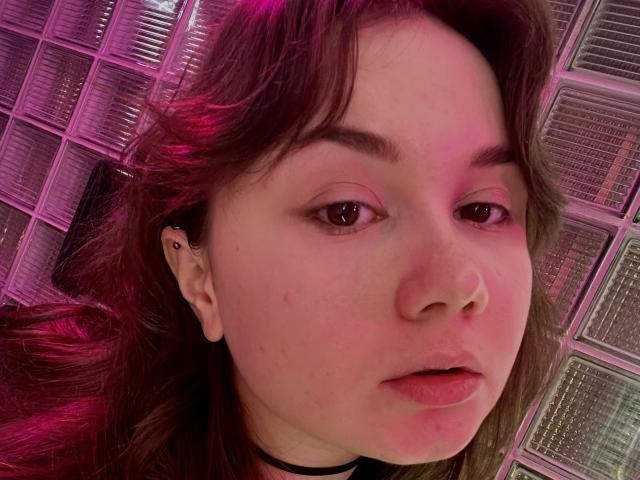 CuteKris has pink-tinted brown hair, brown eyes, small nose, and a black ear piercing. Her face is slightly pink, with a few small acne marks. She's wearing a black choker.