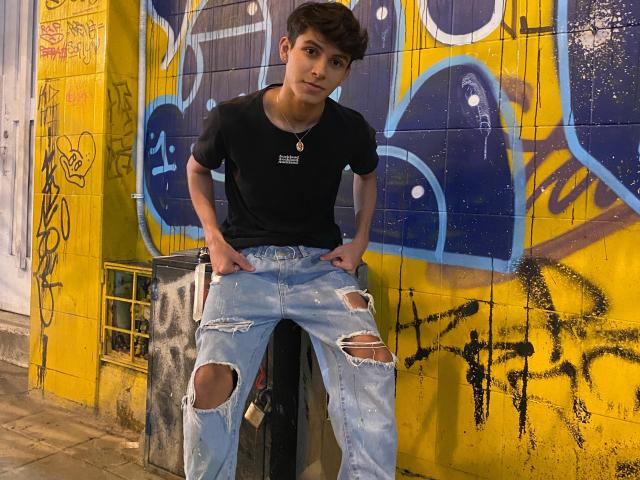Angelitocute with a slim build, short brown hair, black t-shirt, and ripped blue jeans, leaning against a graffiti-covered yellow wall, hands in pockets, looking slightly to the side.