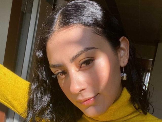 SunnySunset, mit dunklen, welligen Haaren, einem gelben Rollkragenpullover und Diamant-Ohrringen, hat hellbraune Haut, dezentes Make-up und ein leichtes Lächeln. Das Sonnenlicht wirft sanfte Schatten auf ihr Gesicht..