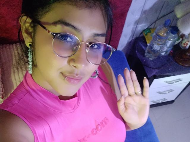 EmiilyJohamson with glasses, pink sleeveless top, earrings, and nose piercing, waving with her right hand, showing a ring, and a lip piercing. Her tits are small.