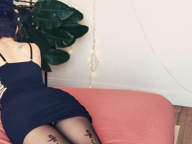 LaPhoenixElo, black dress, black stockings, tattoo on thigh, butt up, blue hair, back tattoo, sitting on pink bed, green plant, string lights, white wall, wooden floor.