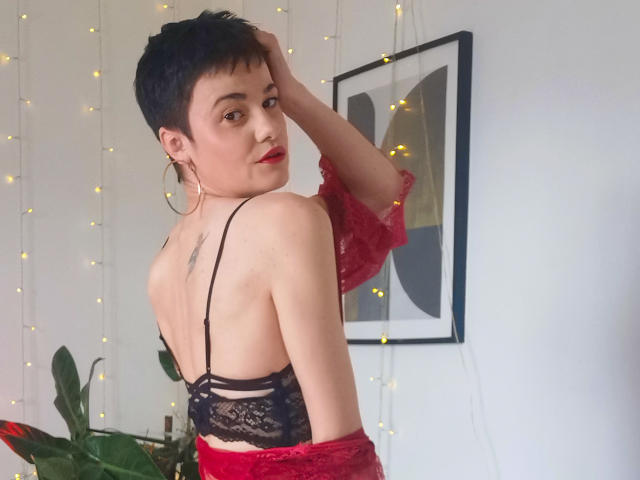LaPhoenixElo with short black hair, red lipstick, hoop earrings, black lace bra, and red lace sleeves, looking over shoulder, showing a small tattoo on her back, in a room.