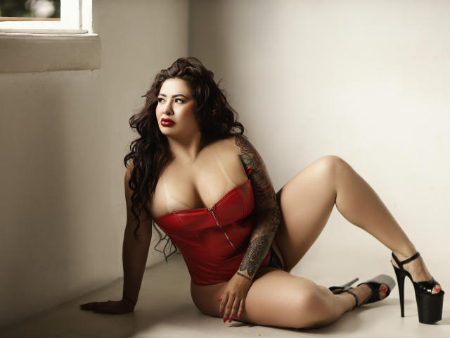 DakotaFranco with big tits, wearing a red zip-up corset, black high heels, tattooed arm, and dark wavy hair, sitting on the floor, looking to the side, sexy.