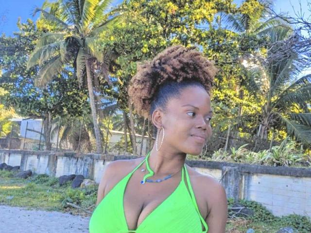 FantineX with curly brown hair in a green halter top, showing her large boobs, wearing hoop earrings and a blue necklace, standing on a sunny beach.