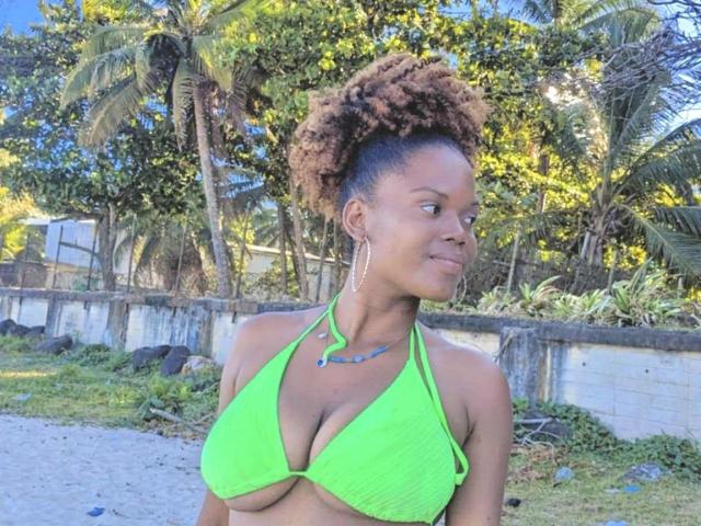 FantineX with brown curly hair in a bright green bikini top, showing her big boobs, standing outside with a confident smile, wearing long earrings and a necklace.