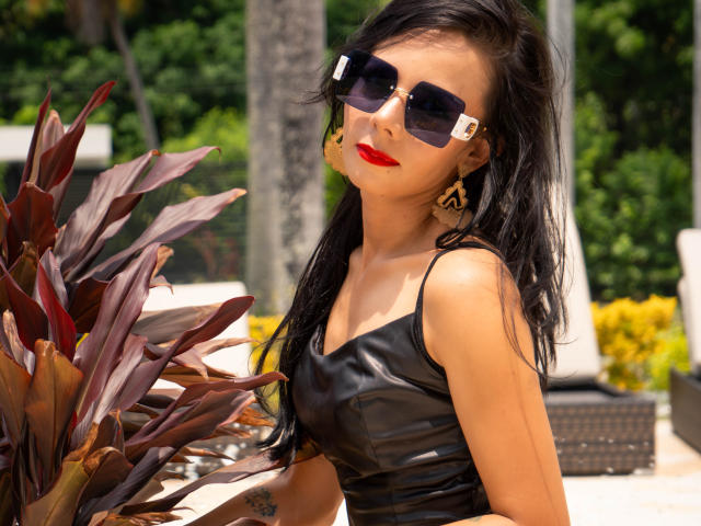 EvelinLuna with dark hair, red lipstick, black leather top, square sunglasses, gold earrings, leaning on plant, sunny, green background, showing a tattoo on her right arm.