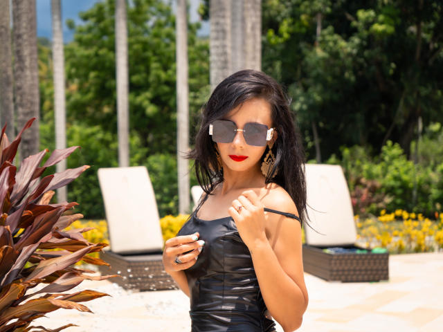 EvelinLuna with black leather dress, red lipstick, and reflective sunglasses, holding dress strap, showing small tits, and black wavy hair, standing by wicker chairs.