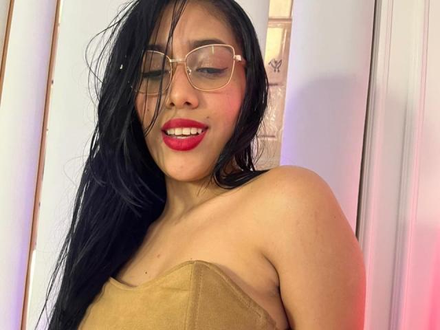 SaraRousse with long black hair, red lipstick, and glasses, wearing a beige towel, smiling with closed eyes, showing her tits. She has a cute face and medium skin.