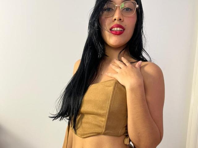 SaraRousse with long black hair, tan strapless top, red lipstick, glasses, and hand on chest, showing her small tits and cute face.
