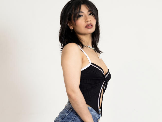SeleneLeon met een gemiddelde borstmaat, in een zwart top met witte accenten en een lage halslijn, en blauwe jeans, staand met de zijkant naar voren, donker haar tot op de schouders, lichte huid, een lichte glimlach, een witte ketting..