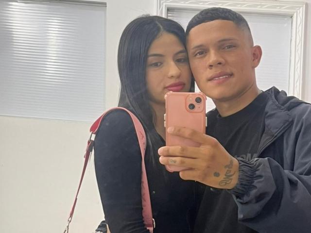 RoseAndDavey, a brunette with long hair and black top, stands next to a tattooed man with a short haircut, taking a selfie with a pink iPhone.