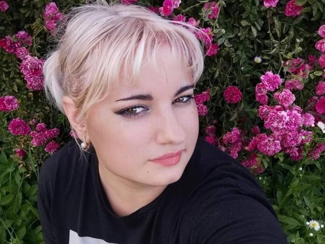 MiLaHottiest has light blonde hair with bangs, black eyeliner, and subtle pink lipstick. She wears a black t-shirt and small earrings. Her skin is fair, and she has a slight, confident smile. The background is filled with pink roses and green leaves, creating a vibrant contrast with her pale hair and black shirt. The image has a natural, candid feel.
