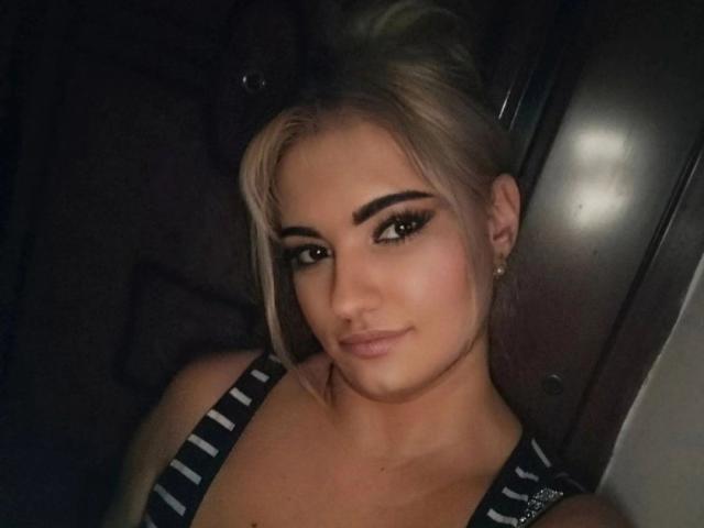 MiLaHottiest has blonde hair in a messy bun, dark eyeliner, and a subtle smile. She's wearing a striped top, showing her tits, with small earrings.