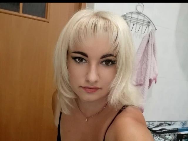 MiLaHottiest has blonde, shoulder-length hair, black eyeliner, and a gold necklace. She's wearing a black top, looking directly at the camera with a subtle smile.