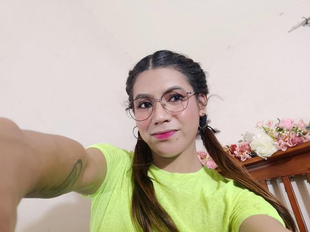 ExpensiveMaria with pigtails, pink lipstick, green shirt, glasses, small nose, tattoo on arm, holding out phone, smiling, pink and white flowers in background.