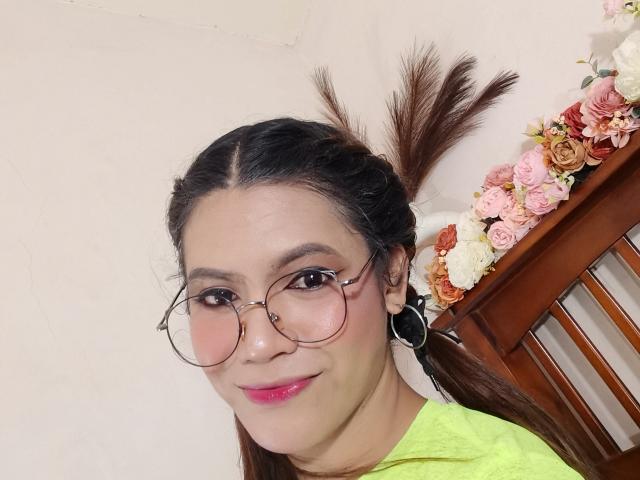 ExpensiveMaria with glasses, pink lipstick, green shirt, black earrings, brown hair in ponytail, smiling at camera, wooden chair, flower arrangement on right.