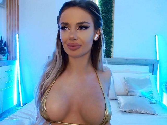 RheaI has long, wavy blonde hair and wears a gold bikini top, exposing large, round breasts. She has full lips, arched eyebrows, and wears white earbuds. The blue lighting in the background highlights her tan skin and makes her appear more prominent. The bed behind her has white pillows and a wooden headboard. The image is a photograph with a focus on RheaI's upper body.