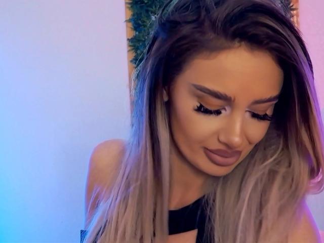 Close-up of RheaI with long, blonde ombre hair, looking downward with eyes closed. She has dark, thick eyebrows, long, curled eyelashes, and dark brown lipstick. She wears a black, strapless top. The background has a blue and purple gradient. RheaI's hair is slightly tousled, and her makeup is bold and defined. The image has a soft, digital filter effect. RheaI's skin is smooth and light. Her expression is subtle and relaxed.