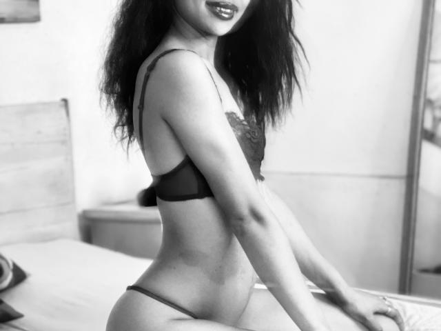 Black and white photo of AubreyLoo, a slim woman with long dark hair, wearing a black lace bra and thong. She's kneeling on a bed, slightly turned to the side, with her right arm resting on the bed. Her lips are darkly painted, and she's looking to the right. The bed has a wooden headboard and a pillow is partially visible. The room is softly lit, creating a contrast between her dark lingerie and lighter skin. Her thong is low-cut, revealing her hip bones. The image focuses on her upper body and face, with a slight blur on the background.