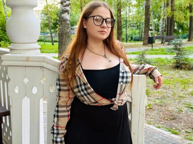 AnnieWow-hot with glasses, long brown hair, plaid shirt tied at the front, black dress, and necklace, leaning on white railing in park, looking slightly to the side.