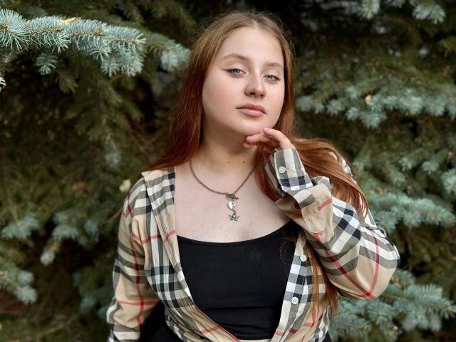 Annie, een aantrekkelijke vrouw met lang rood haar, draagt een geruite blouse over een zwarte top. Ze heeft haar hand in de buurt van haar mond, draagt een ketting met een ster en heeft een rustige uitdrukking..