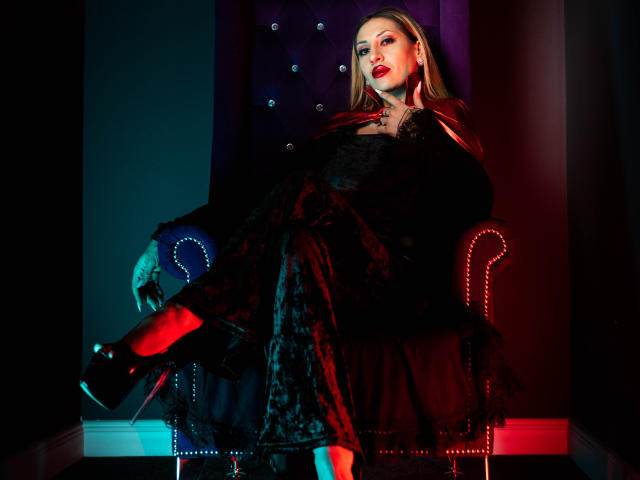 VickiSpices with blonde hair, red lipstick, and black lace dress, sitting in a dark chair, legs crossed, wearing high heels, and a red cape draped over her shoulders.