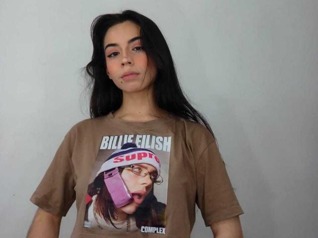 KatherinJohns with long black hair, brown t-shirt with a Supercross graphic, and a small lip piercing, looking at the camera with a slight smile.